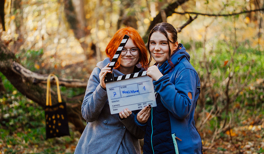 Zwei junge Mädchen stehen neben einander in einem wald. Sie halten gemeinsam eine Filmklappe in den Händen. 